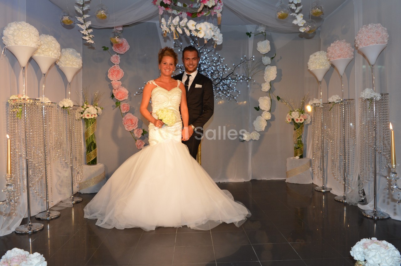 Un couple en tenue de mariage pose devant une décoration florale élégante dans une salle ornée de voilages blancs.