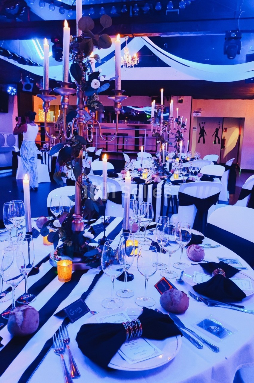 Salle de réception décorée avec des chandeliers, nappes blanches et noires, verres et bougies sur les tables.