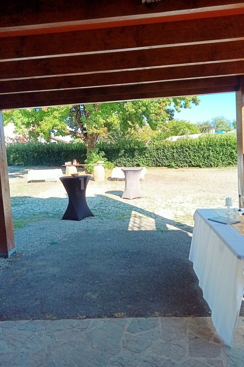 Vue depuis un espace couvert sur une cour extérieure avec tables hautes noires et un arbre en arrière-plan.