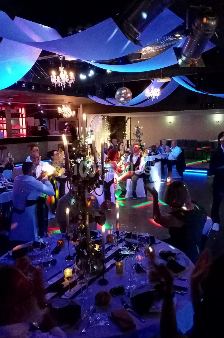 Salle de réception décorée avec des guirlandes et des lumières colorées, des invités assis et dansant.