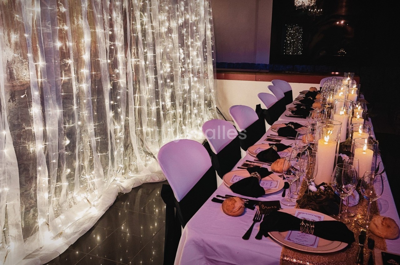 Table élégamment dressée avec nappes blanches, bougies, serviettes noires et guirlandes lumineuses en arrière-plan.