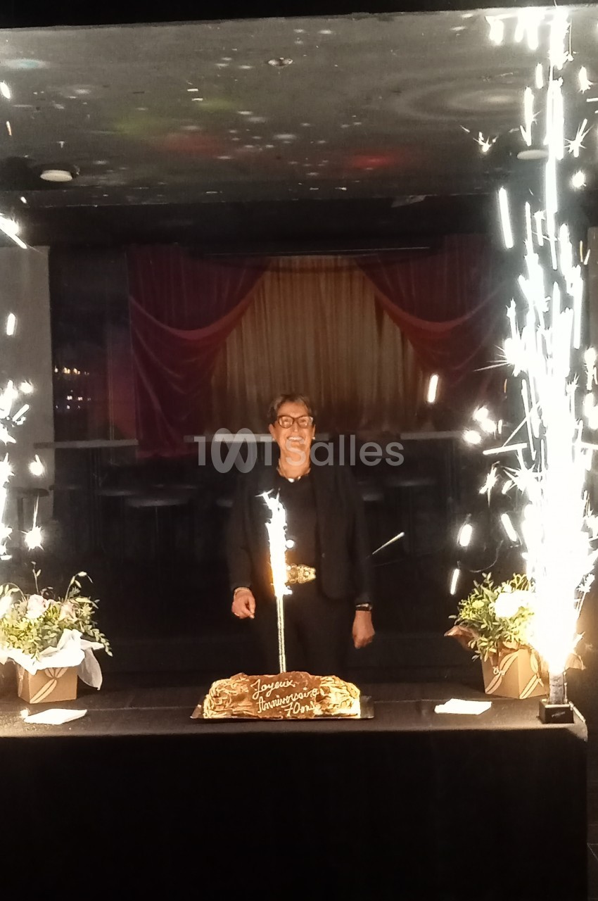 Une personne souriante debout derrière un gâteau d'anniversaire entouré de fontaines lumineuses et de décorations.