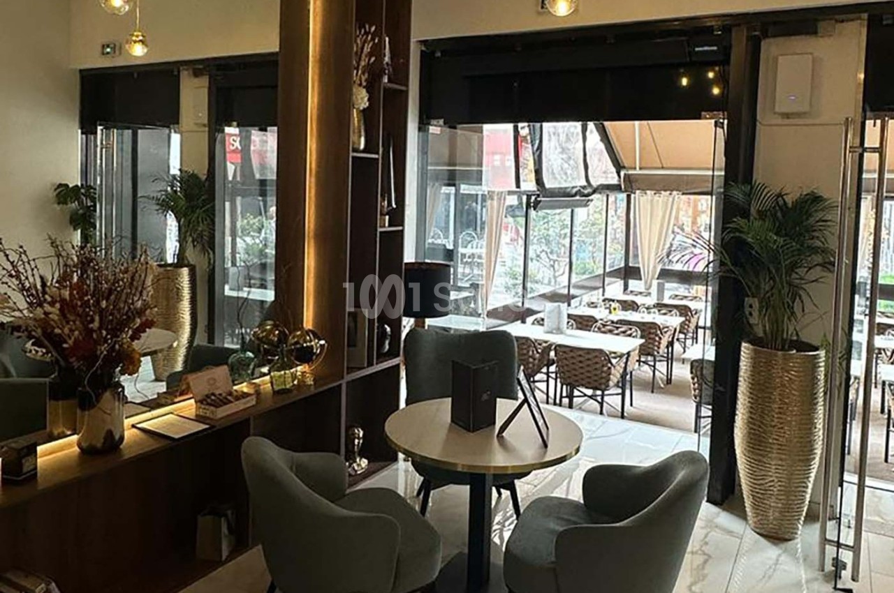 Intérieur d'un café moderne avec tables, chaises rembourrées, plantes décoratives et vue sur une terrasse extérieure.