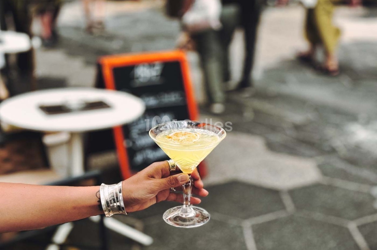 Une main tenant un verre de cocktail jaune avec une rondelle de citron, en terrasse avec des tables et un tableau en arrière…