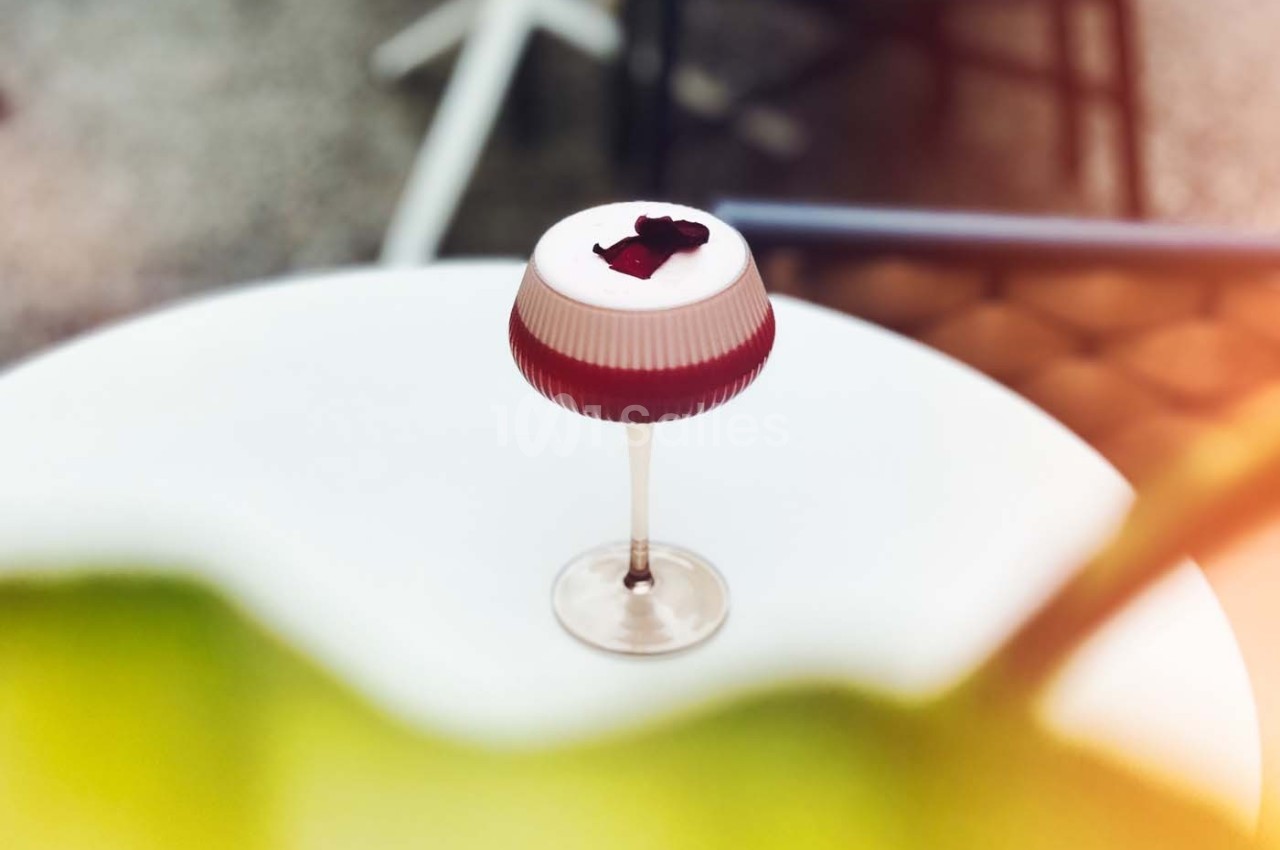 Verre à cocktail avec mousse blanche et pétales rouges, posé sur une table blanche dans un environnement lumineux.
