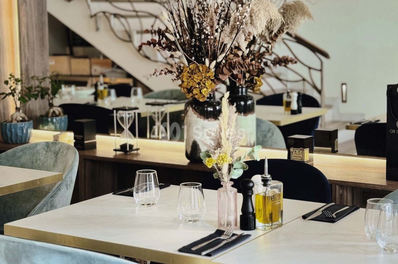 Table dressée avec des verres, couverts et condiments, entourée de chaises, dans un restaurant au décor élégant avec plantes.