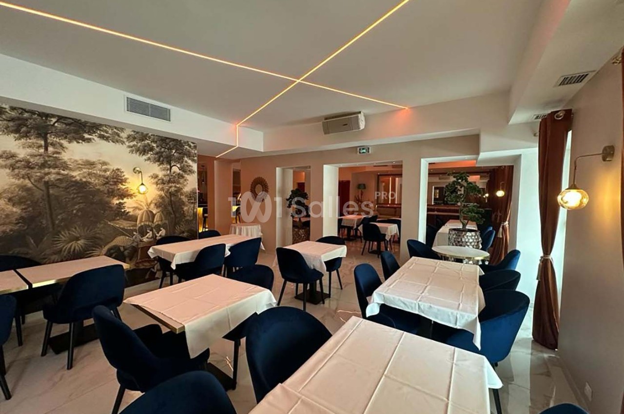 Salle de restaurant lumineuse avec tables dressées, chaises bleues et décoration murale végétale.