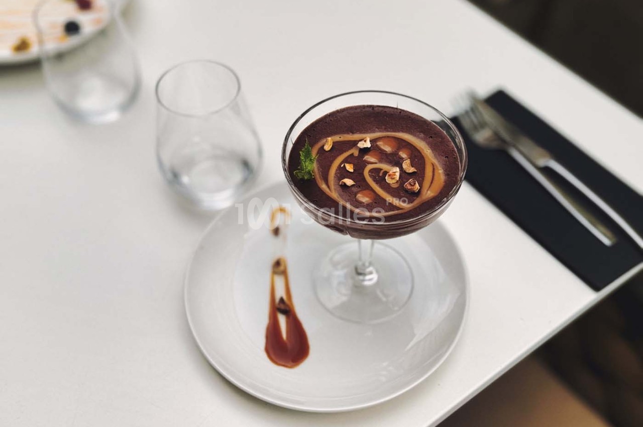 Dessert au chocolat dans une coupe, décoré de noisettes et d'un filet de sauce, servi sur une assiette blanche.