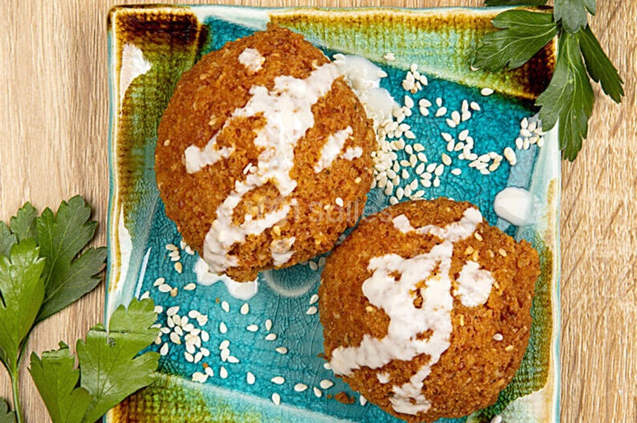 Deux falafels nappés de sauce blanche et parsemés de graines de sésame, présentés sur une assiette colorée avec du persil.