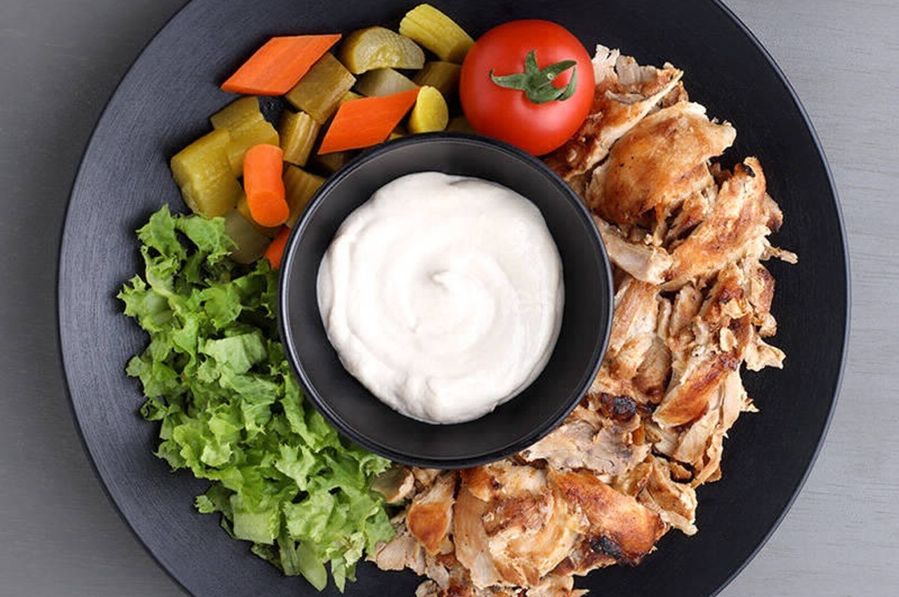 Assiette contenant du poulet grillé, sauce blanche, salade verte, légumes marinés et une tomate cerise.