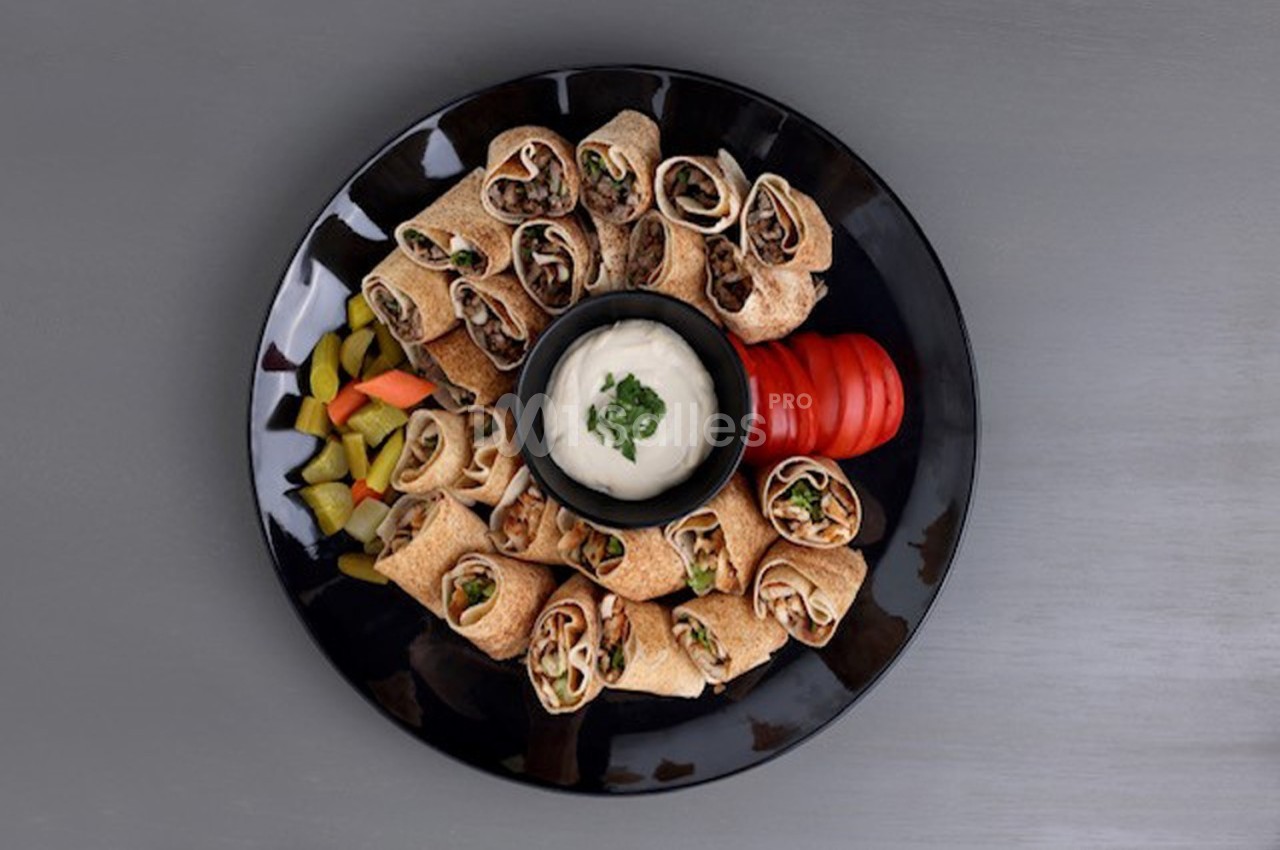 Assiette noire contenant des wraps garnis, accompagnée de légumes, de tranches de tomate et d'une sauce blanche au centre.