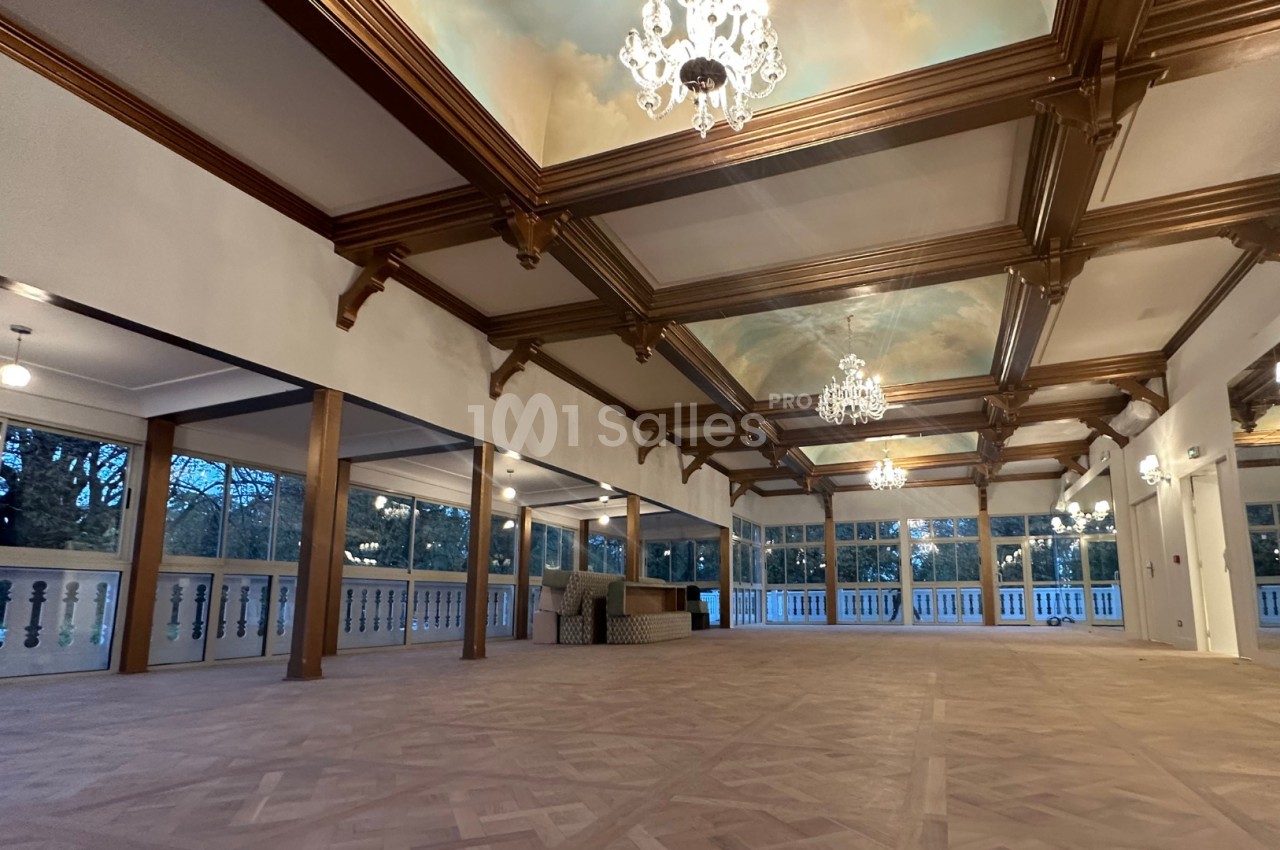 Salle spacieuse avec parquet en bois, plafond à caissons décoré de fresques et lustres, entourée de grandes fenêtres.