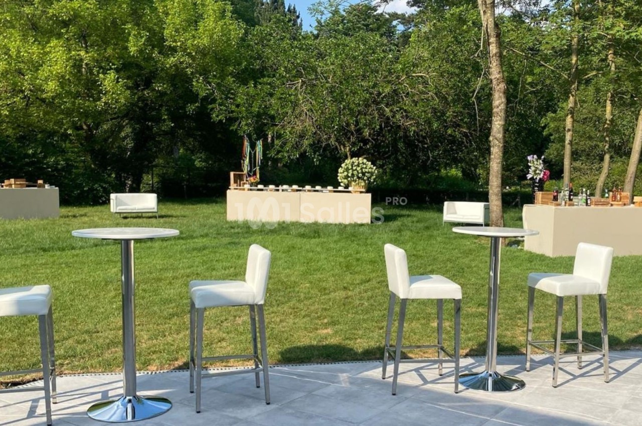 Tables hautes et chaises blanches disposées sur une terrasse, avec un jardin verdoyant et des buffets en arrière-plan.