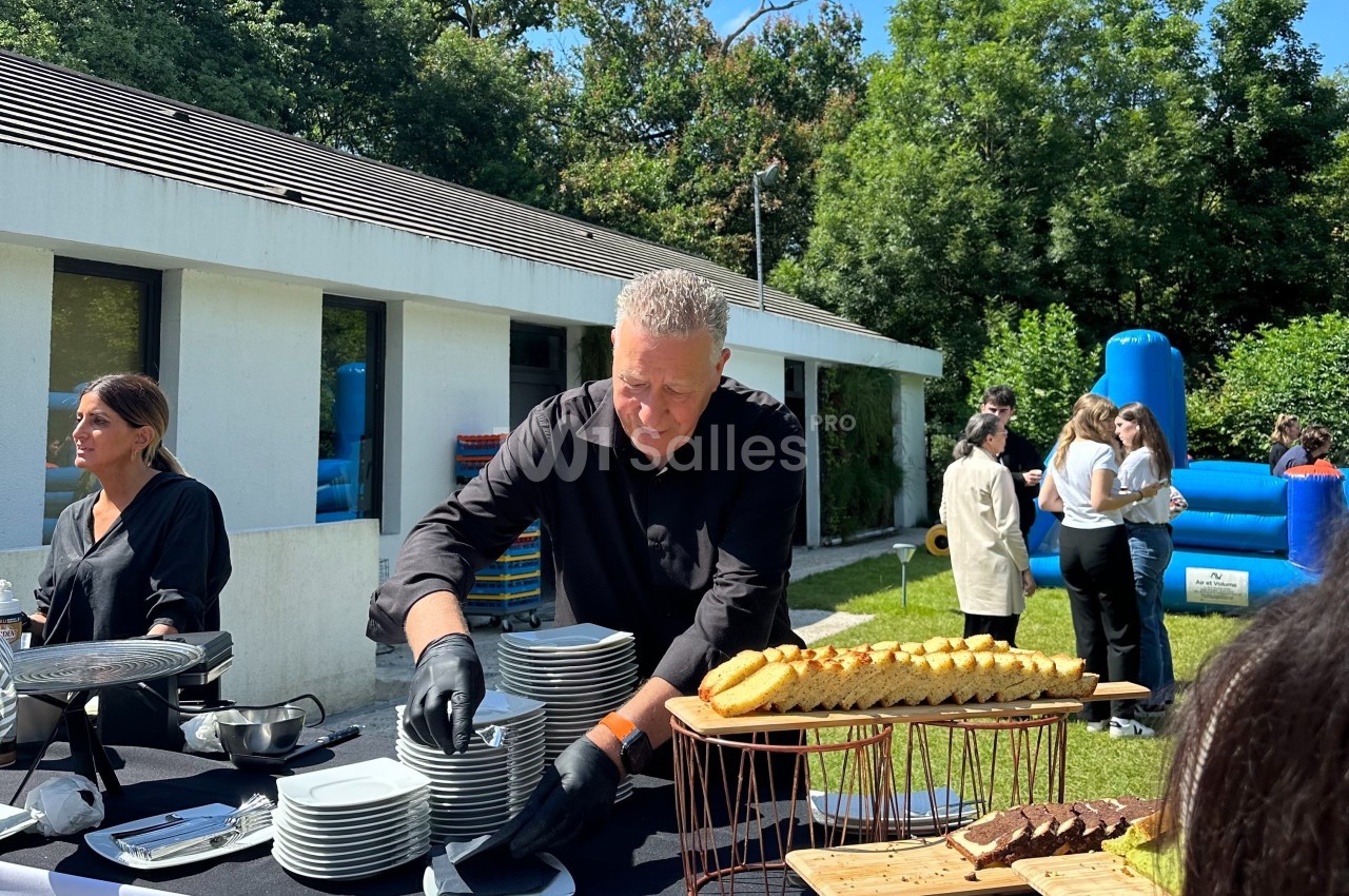 Un homme sert des assiettes sur une table de buffet en extérieur, avec des invités et un château gonflable en arrière-plan.