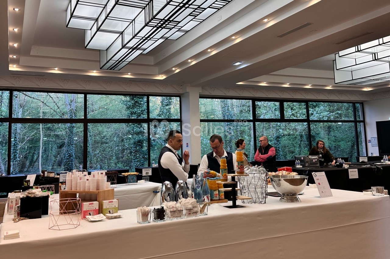 Buffet avec boissons, snacks et serveurs dans une salle lumineuse avec de grandes baies vitrées donnant sur un espace arboré.