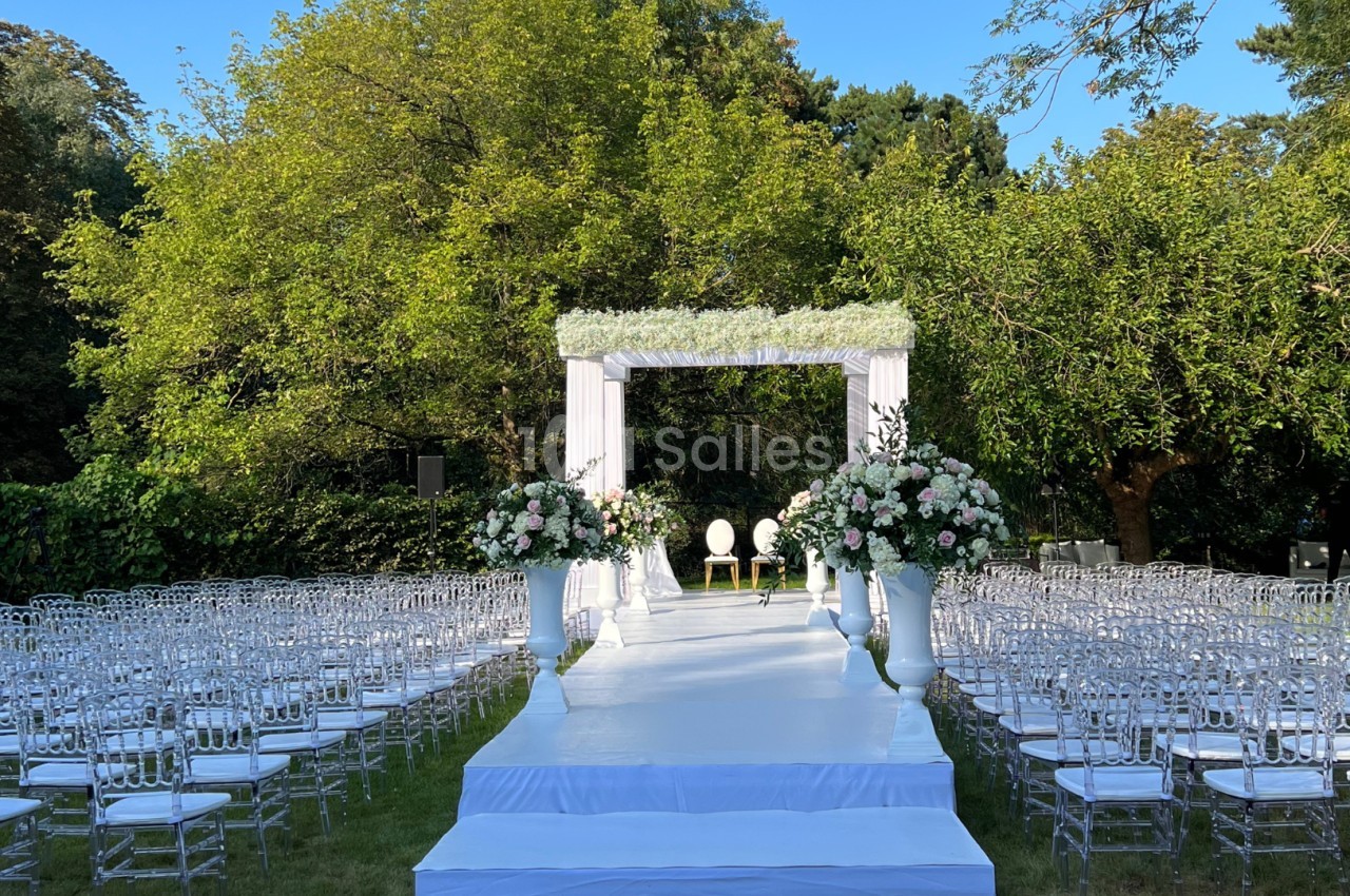 Allée centrale menant à une arche de mariage décorée de fleurs blanches et roses, entourée de chaises transparentes en…