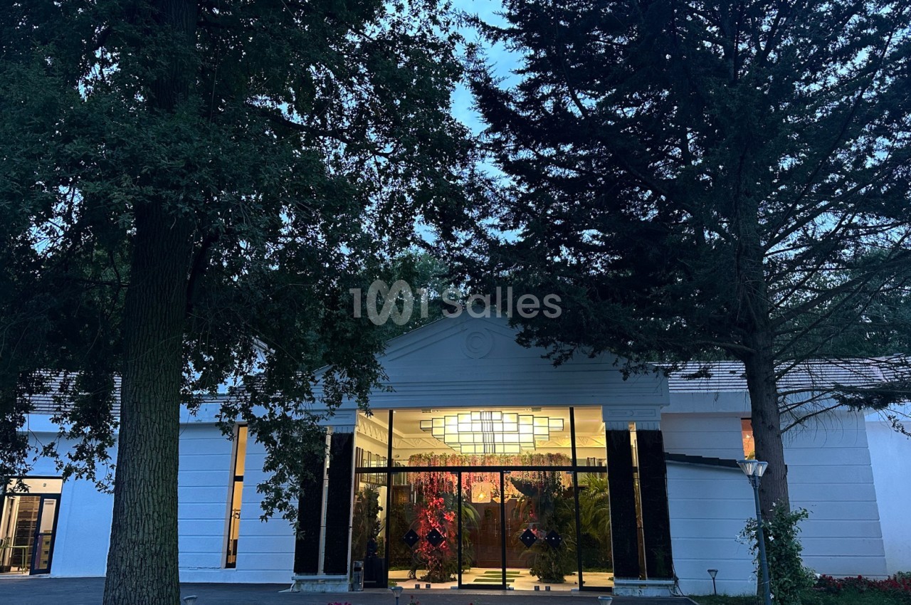 Entrée d'un bâtiment blanc entouré d'arbres, avec des portes vitrées éclairées et un lustre visible à l'intérieur.
