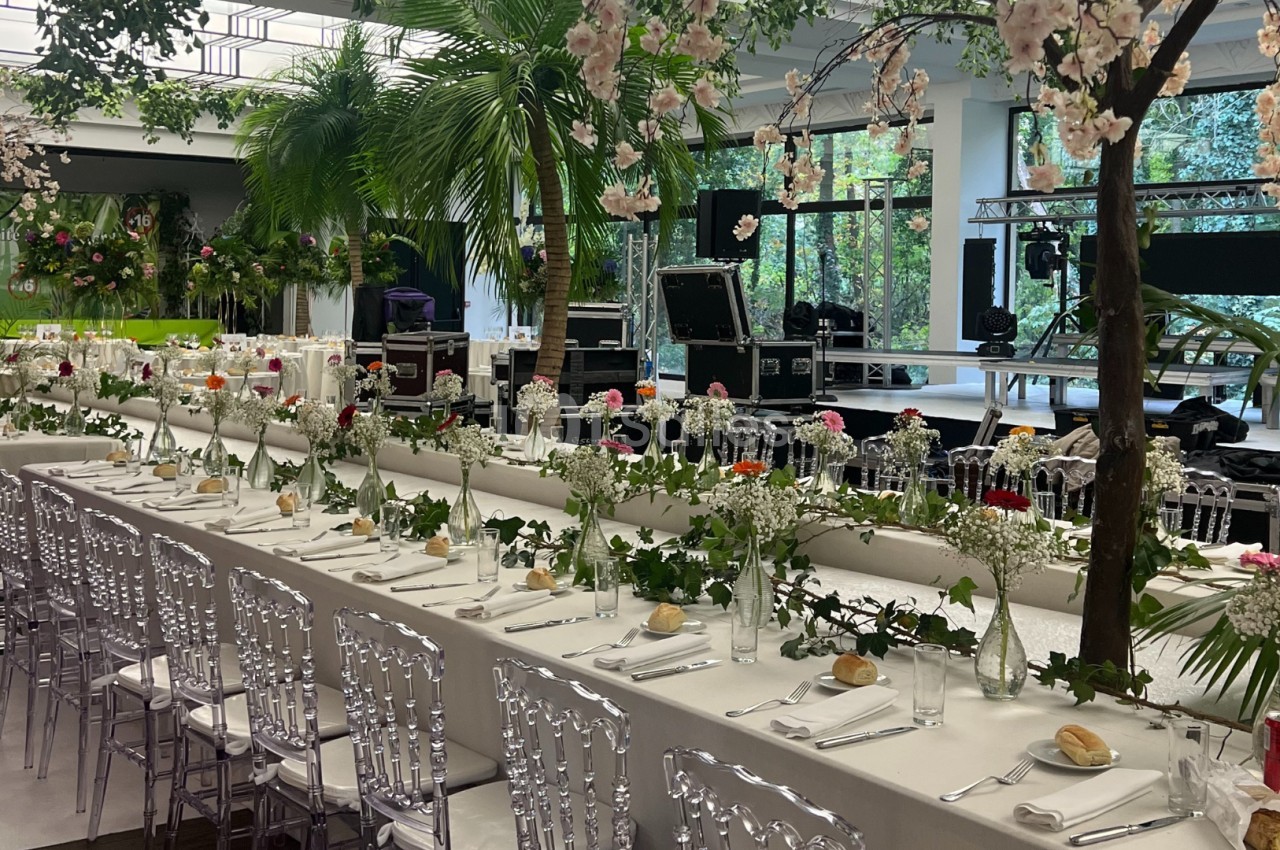 Grande table décorée de fleurs blanches et roses, entourée de chaises transparentes, dans une salle lumineuse avec plantes.