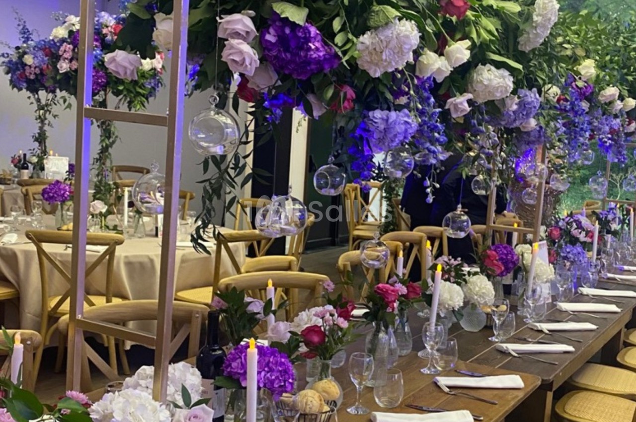 Table décorée pour un événement avec des fleurs suspendues, des bougies et une vaisselle élégante dans une salle lumineuse.