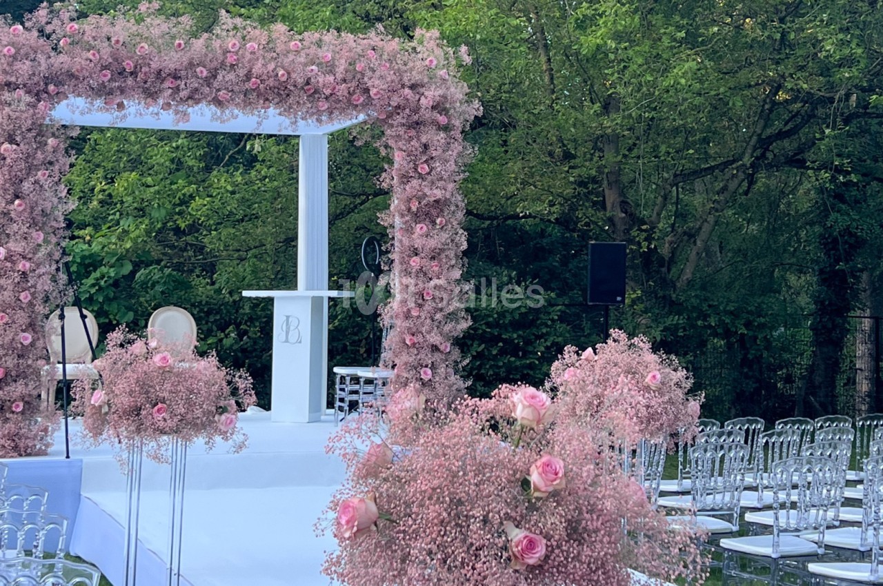 Arche de cérémonie ornée de fleurs roses, entourée de chaises transparentes, dans un jardin verdoyant.