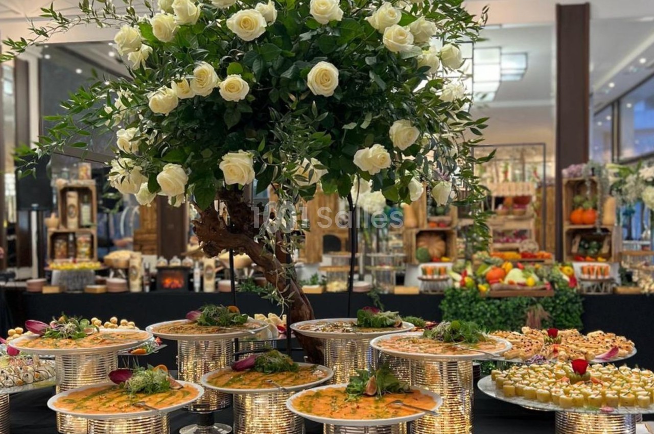 Buffet élégant avec plats variés sur des présentoirs argentés, décoré d'un grand bouquet de roses blanches.
