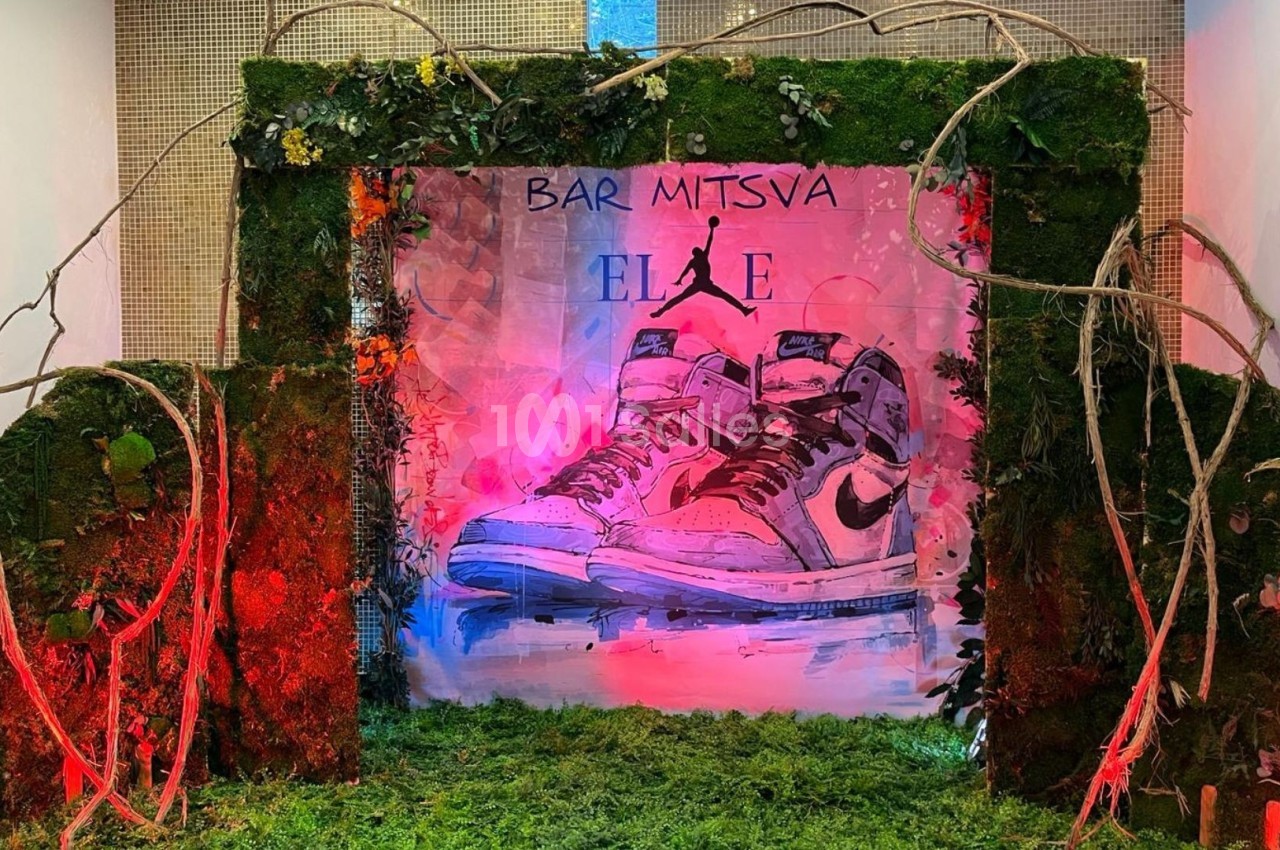Décor de bar-mitsva avec des baskets illustrées, entouré de mousse, branches et éclairages colorés.