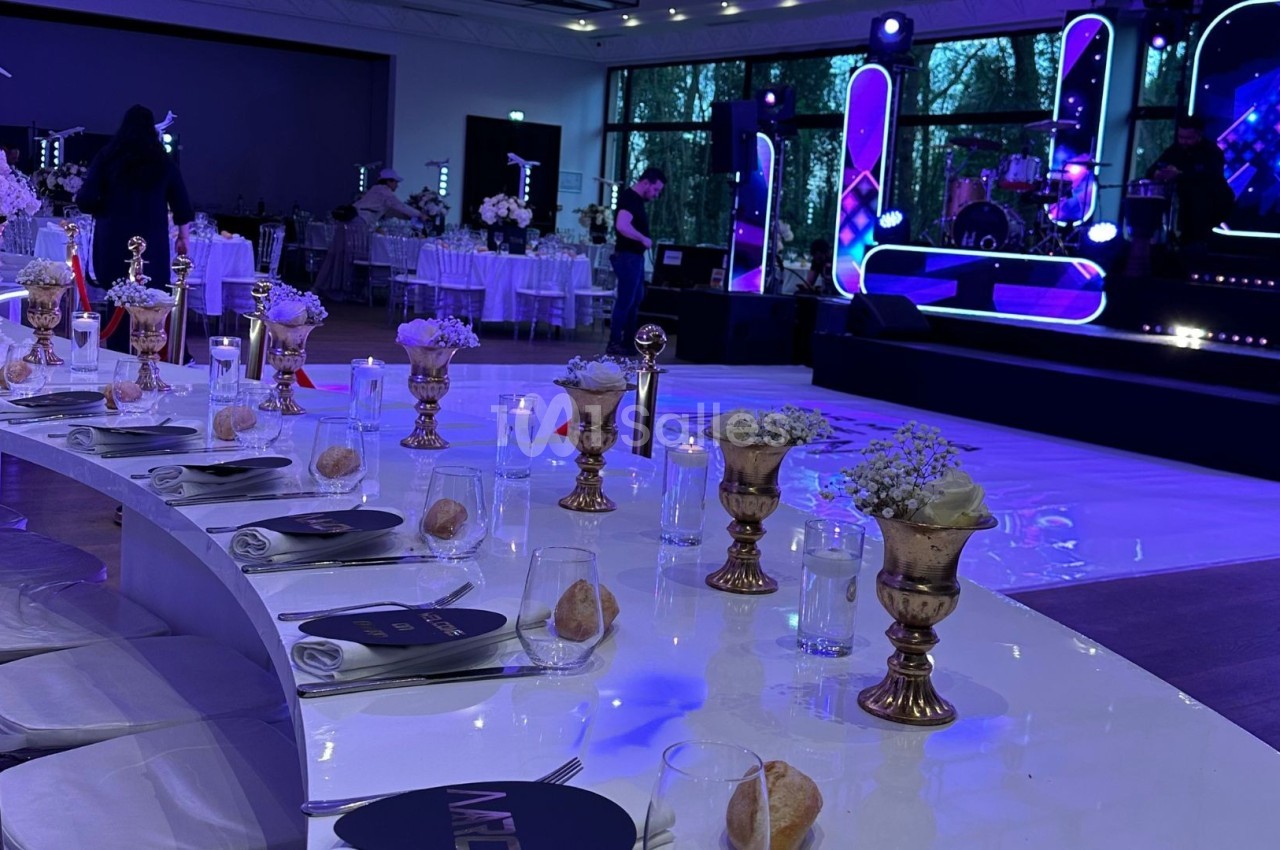 Table décorée avec des vases dorés et des fleurs blanches, face à une scène illuminée dans une salle de réception.
