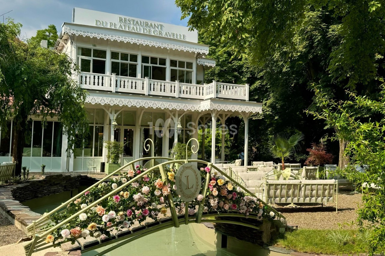 Façade d'un restaurant élégant entouré de verdure, avec un pont fleuri au premier plan.