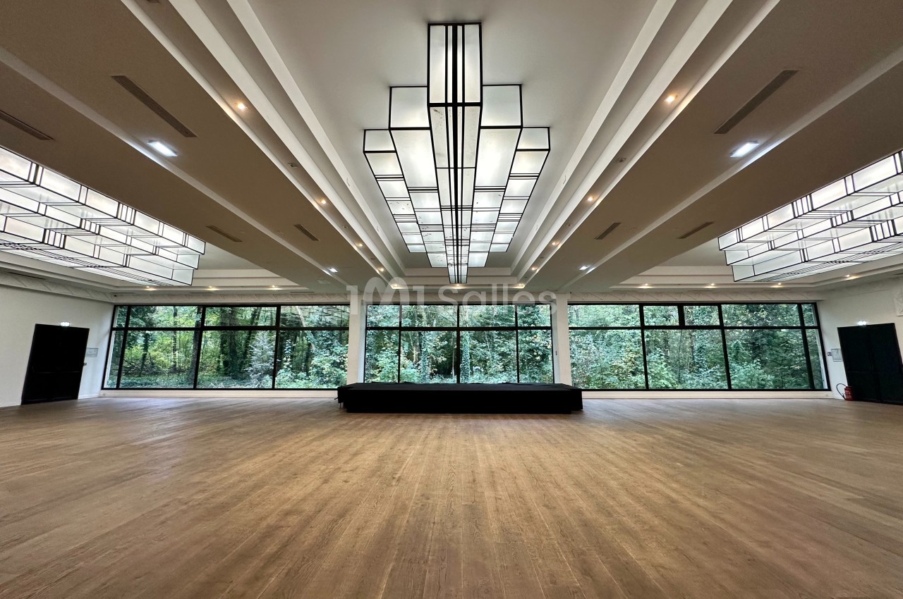 Salle spacieuse avec parquet clair, grandes baies vitrées donnant sur des arbres et plafonniers géométriques modernes.