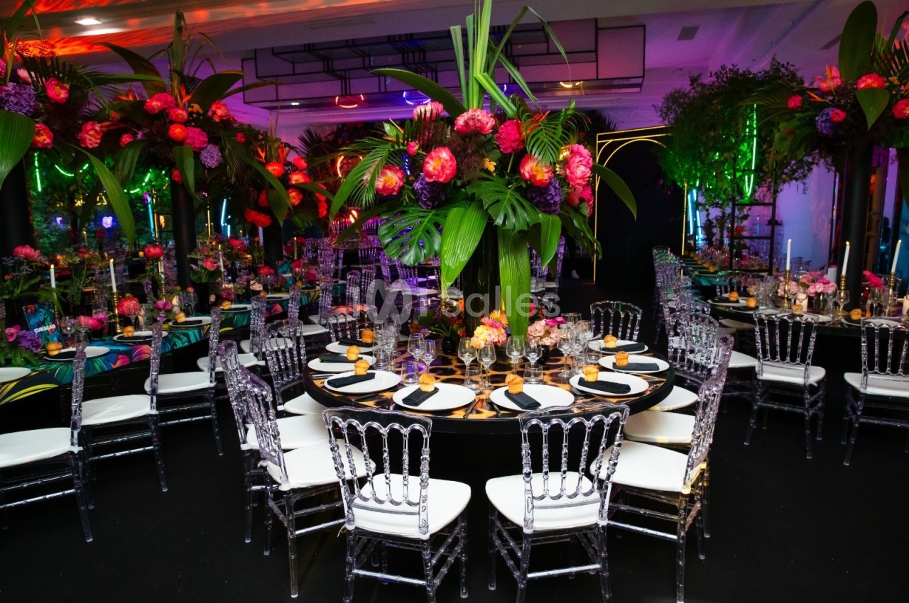 Salle de réception décorée avec des tables rondes, des chaises transparentes et des arrangements floraux colorés.
