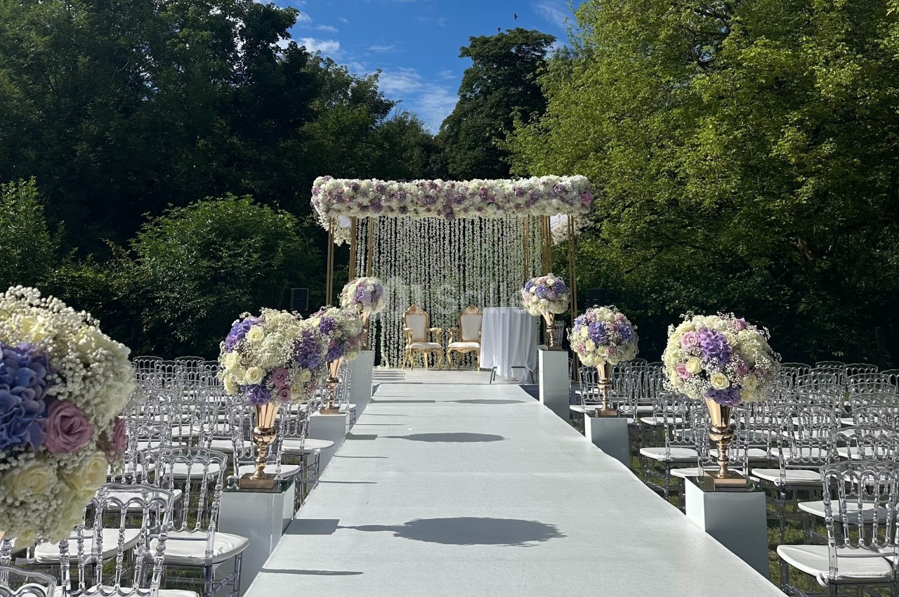 Allée décorée de fleurs menant à une arche florale blanche et dorée pour une cérémonie en plein air.