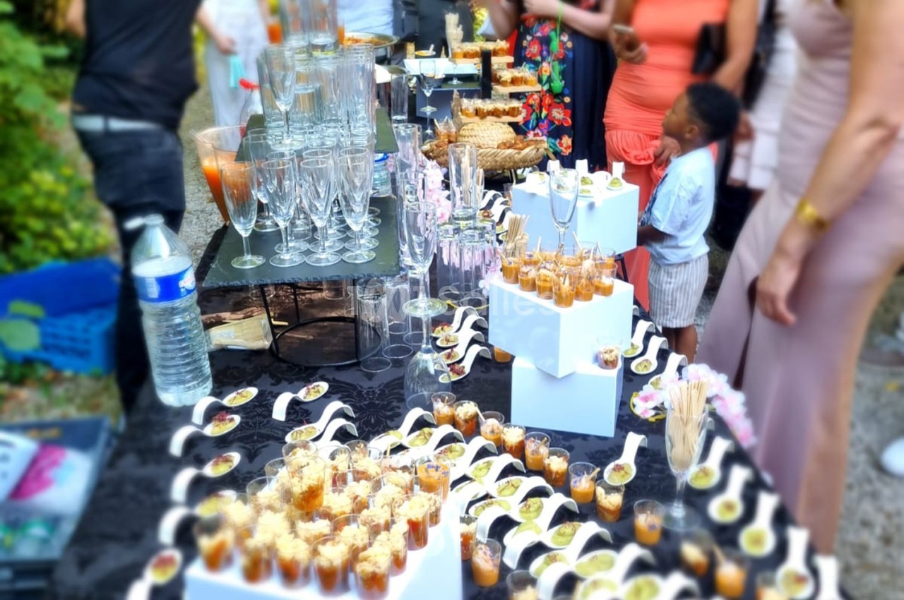 Buffet en extérieur avec verrines, amuse-bouches et verres disposés sur une table noire, entourée de convives.