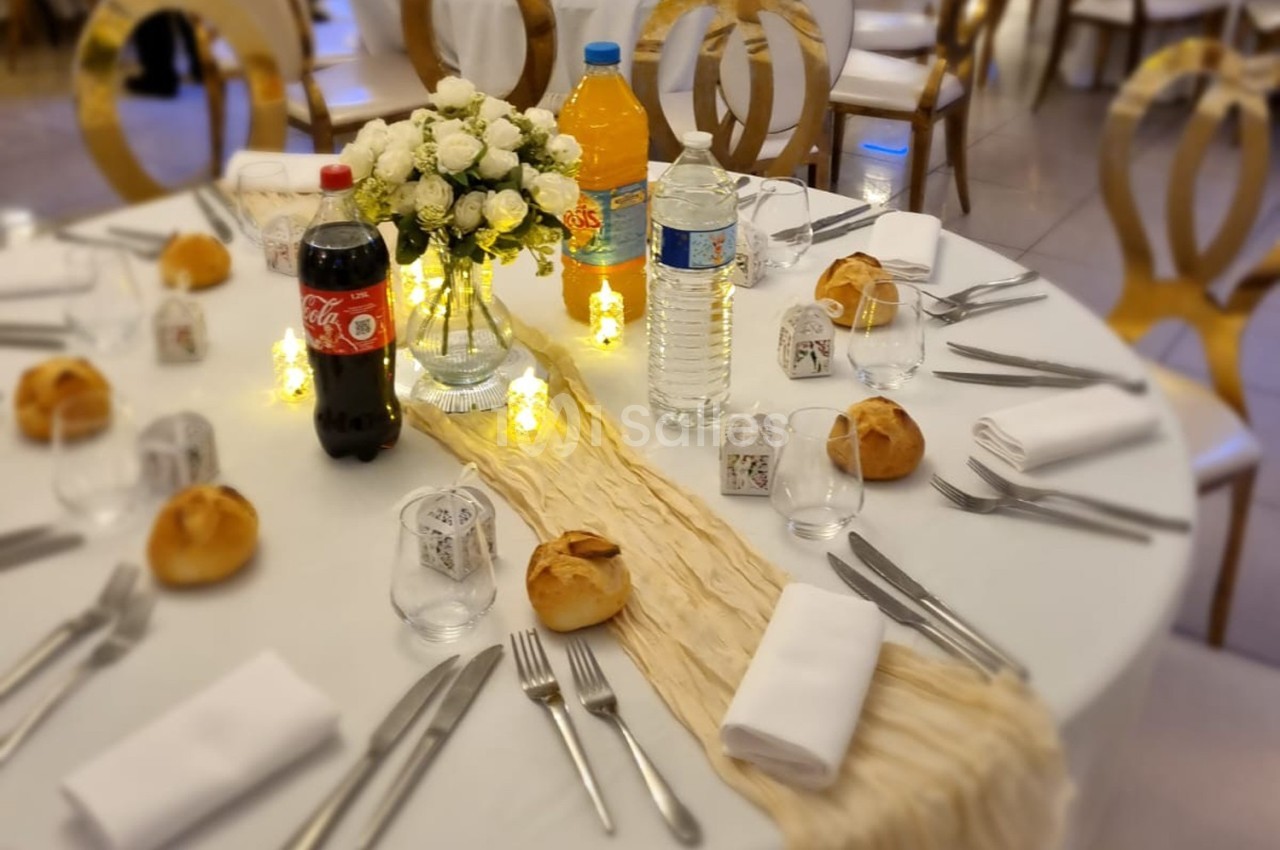 Table dressée pour un repas avec nappes blanches, couverts, pains, bouteilles de boissons et décoration florale centrale.