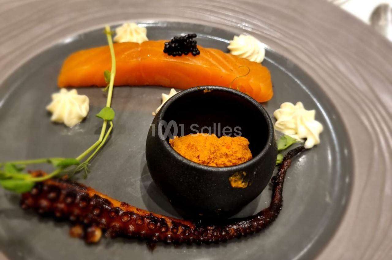 Assiette gastronomique avec saumon fumé, tentacule de poulpe grillé, épices dans un bol noir et décorations végétales.