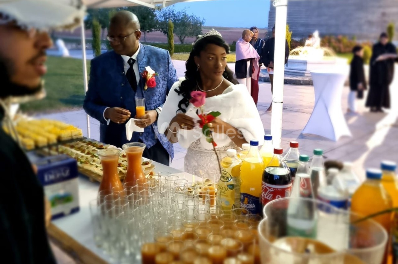 Un couple en tenue de mariage se sert à un buffet en plein air, entouré de boissons et de verrines.