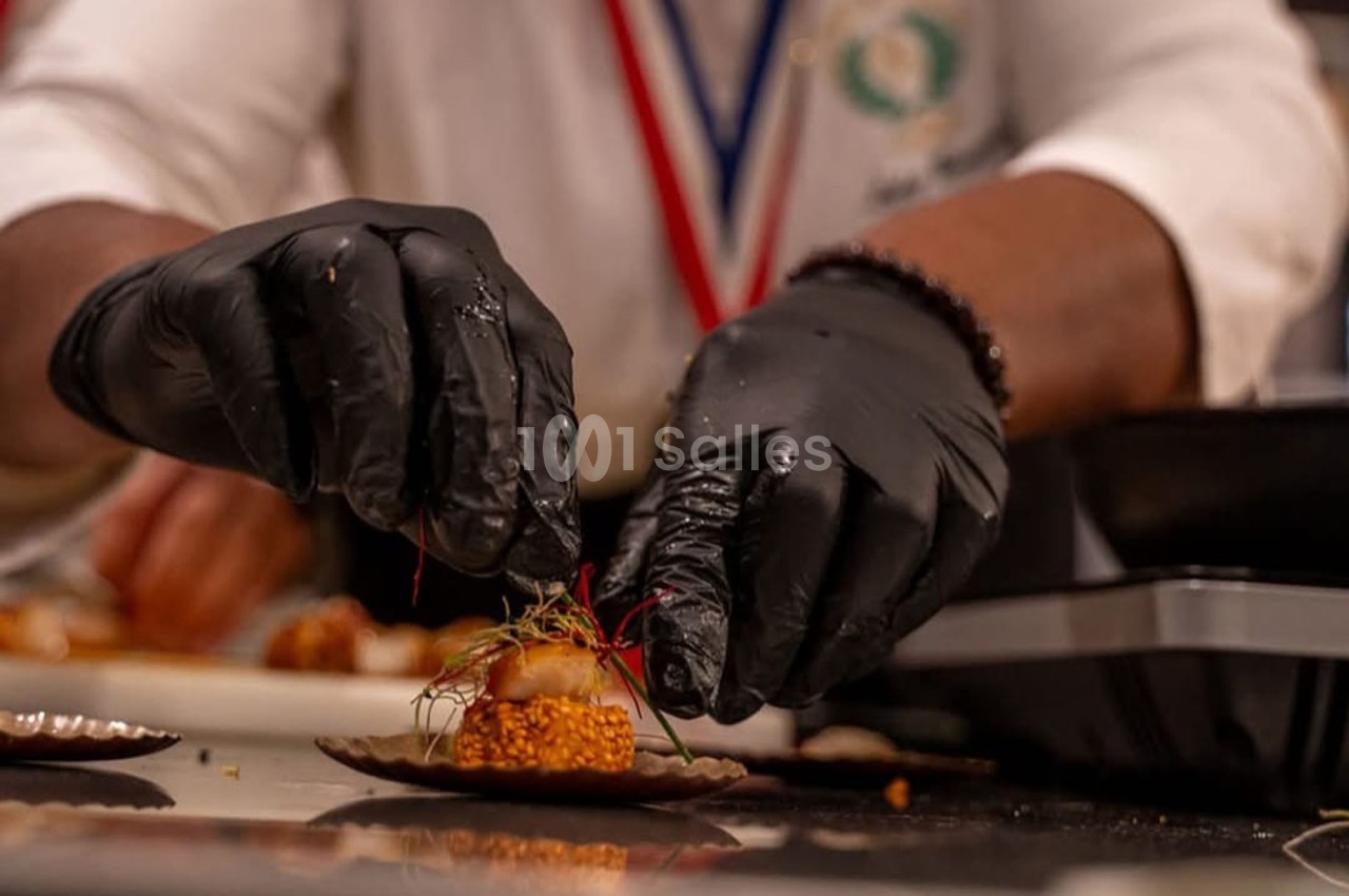 Un chef en gants noirs décore un plat avec des herbes fines dans une cuisine professionnelle.