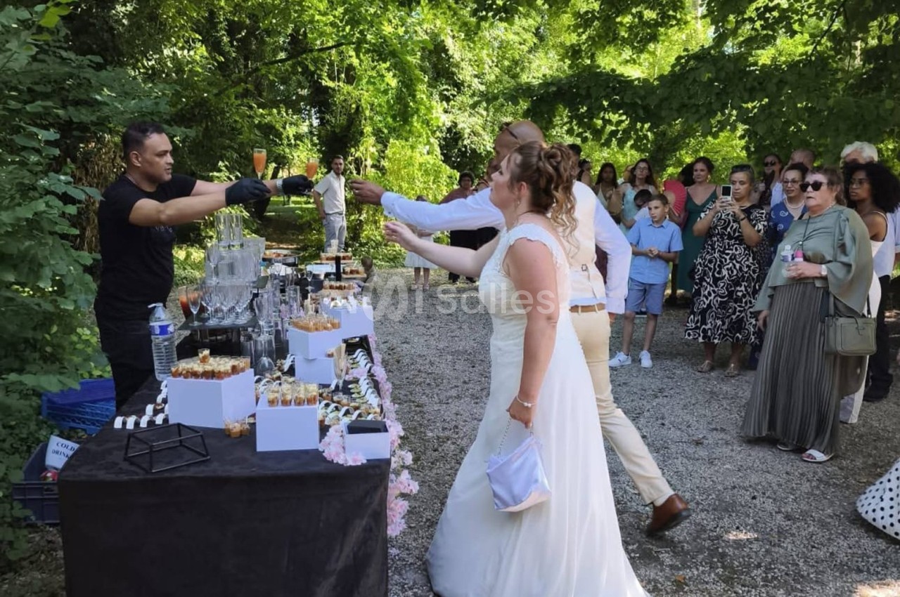 Un couple en tenue de mariage trinque avec un serveur devant une table de buffet en extérieur, entouré d'invités.