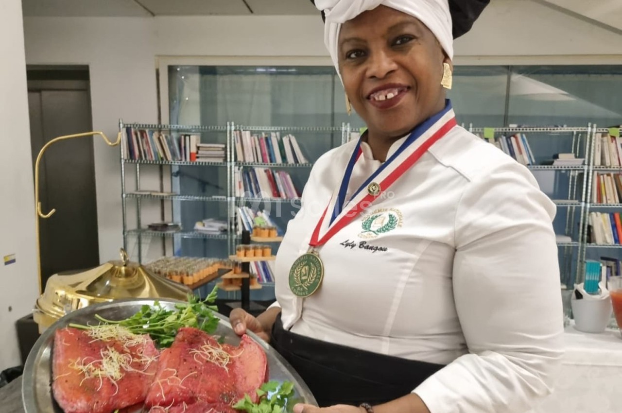 Une cheffe en tenue professionnelle présente un plateau de viande garnie de pousses, dans une cuisine avec des étagères en…