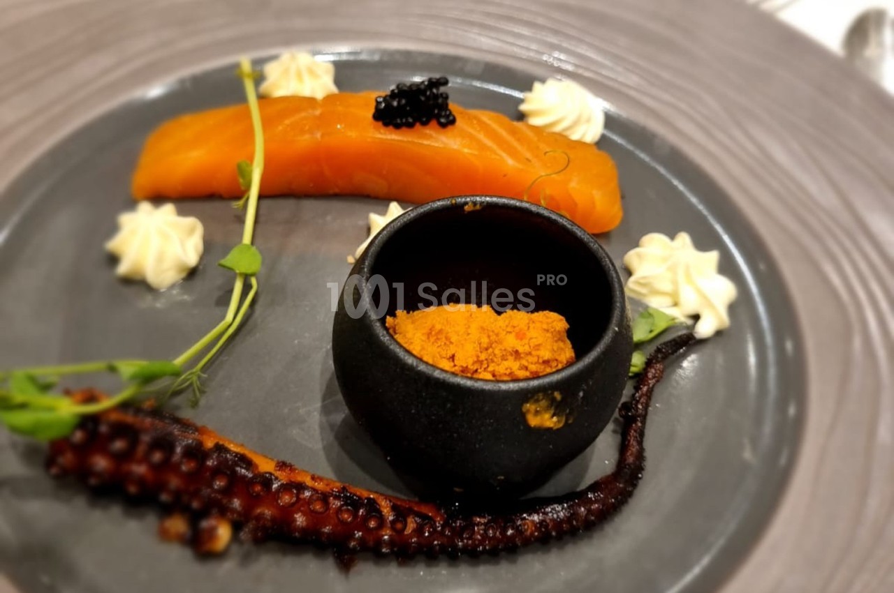 Assiette gastronomique avec saumon fumé, tentacule de poulpe grillé, épices dans un bol noir et décorations végétales.