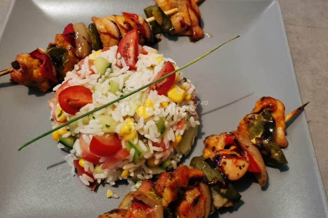 Assiette avec brochettes de poulet grillé et salade de riz garnie de légumes frais, servie sur une assiette grise.