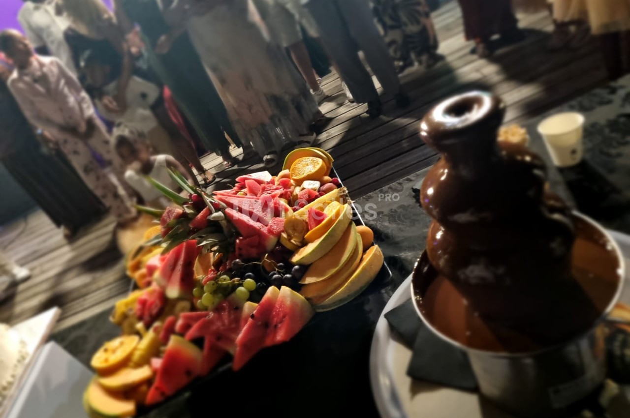 Plateau de fruits variés et fontaine de chocolat au premier plan, avec des personnes en arrière-plan flou.