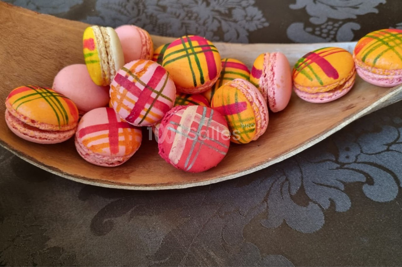 Macarons colorés avec motifs à carreaux disposés dans une cuillère en bois sur une nappe à motifs floraux.