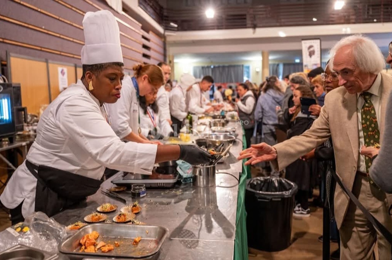 Une cheffe sert des plats à un homme lors d'un événement culinaire dans une salle bondée.