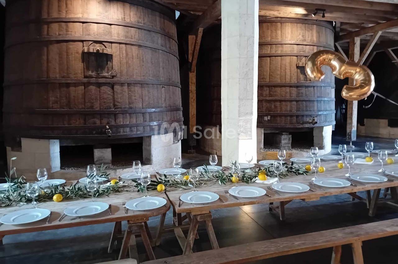 Table en bois dressée avec des assiettes, verres et décoration végétale, devant de grands tonneaux en bois.