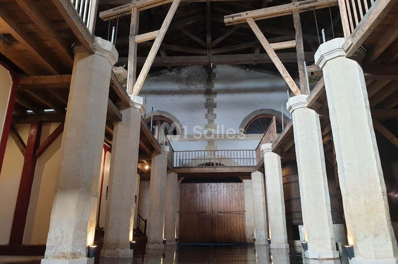 Intérieur d'un bâtiment avec des colonnes en pierre, une charpente en bois apparente et un sol réfléchissant.