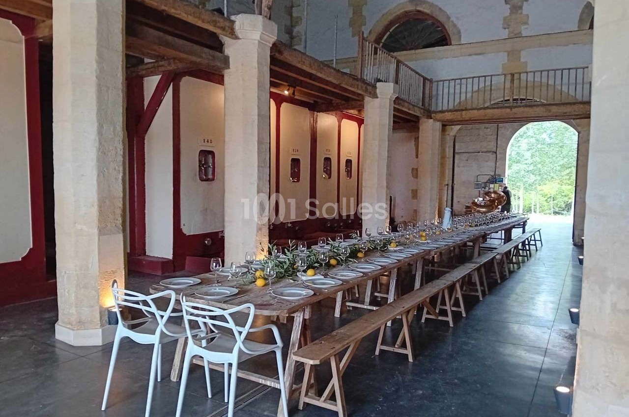 Grande table en bois dressée pour un repas dans une salle aux murs en pierre avec des cuves de vin en arrière-plan.