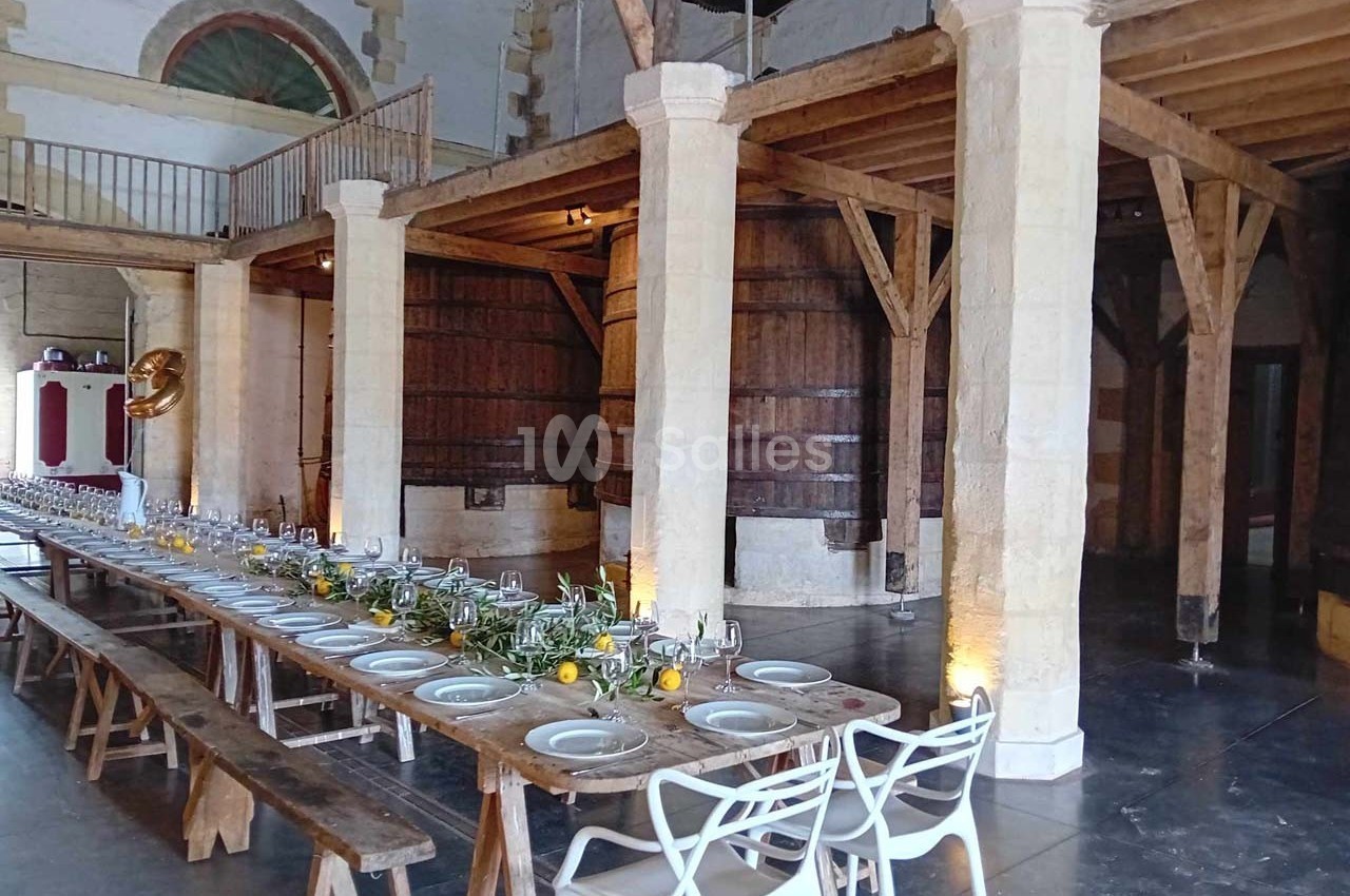 Grande salle rustique avec longues tables en bois dressées, décorées de verdure et de citrons, entourées de tonneaux en…