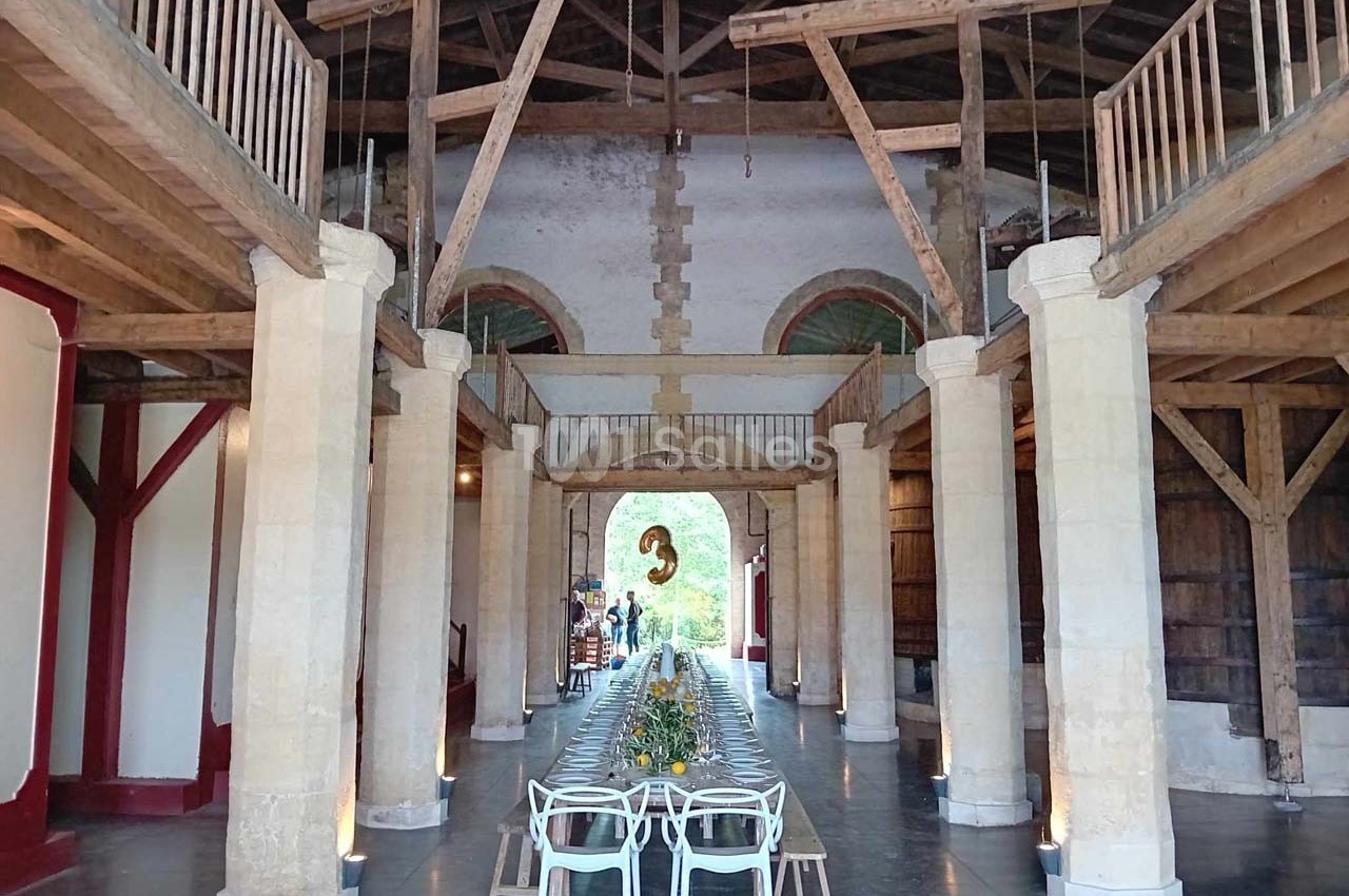 Grande salle avec charpente en bois apparente, table longue dressée au centre et décorations lumineuses.