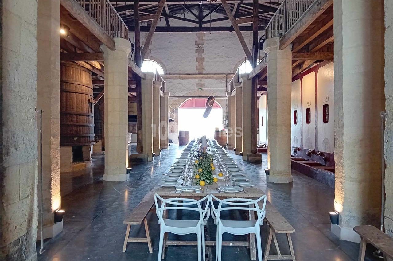Grande salle avec une longue table dressée pour un repas, entourée de bancs et de chaises, dans un cadre rustique avec…