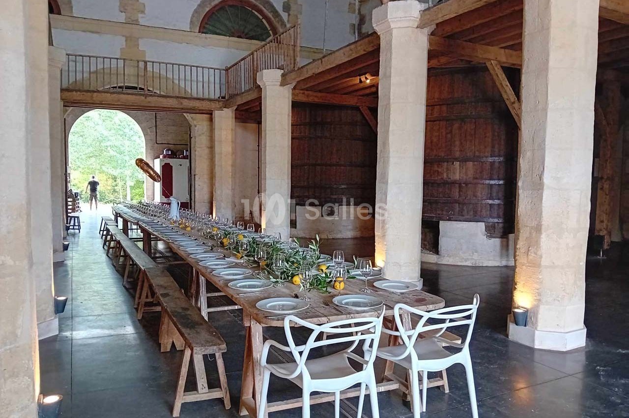 Grande table en bois dressée avec vaisselle et décorations florales dans une salle rustique avec poutres et tonneaux.